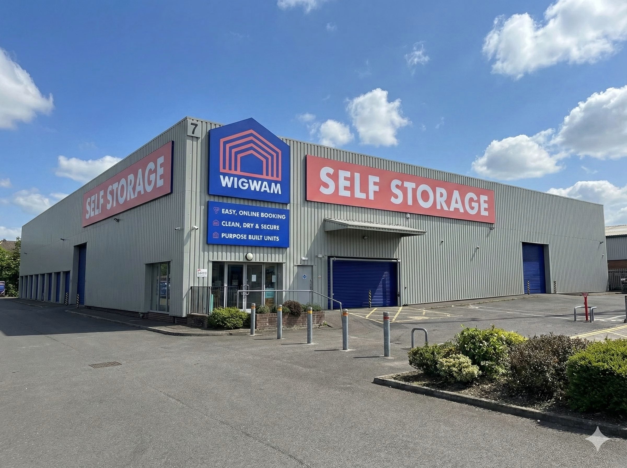 A large Wigwam Self Storage facility with grey cladding, bold red and blue signage and multiple roller-shutter doors, photographed on a clear day with a blue sky. The site features a clean forecourt, customer entrance and purpose-built storage units, representing a modern self-storage operation.